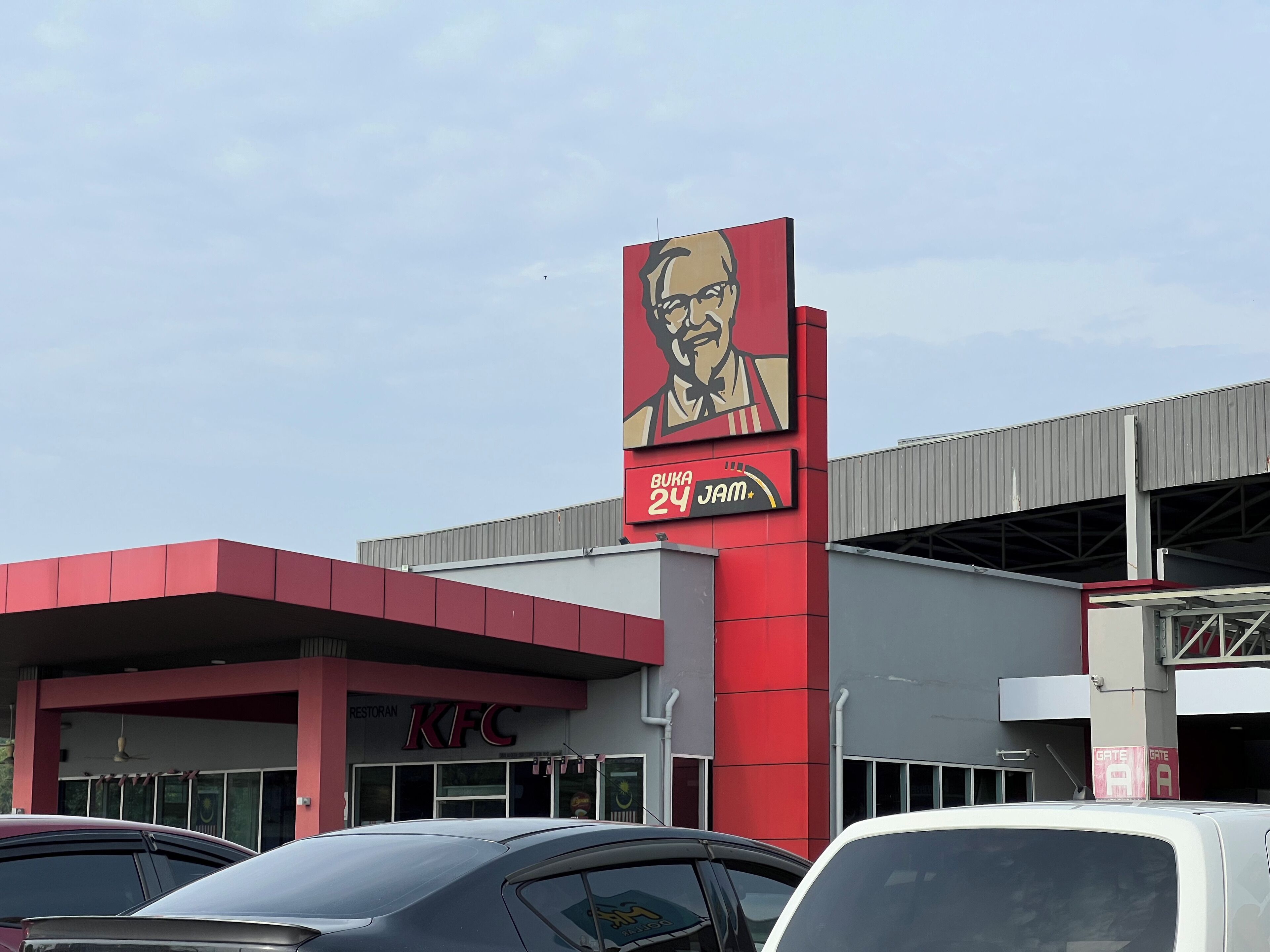 Um restaurante KFC com esquema de cores vermelho e branco marcante, um grande outdoor anunciando 'Buka 24 Jam' e um estacionamento com vários carros.