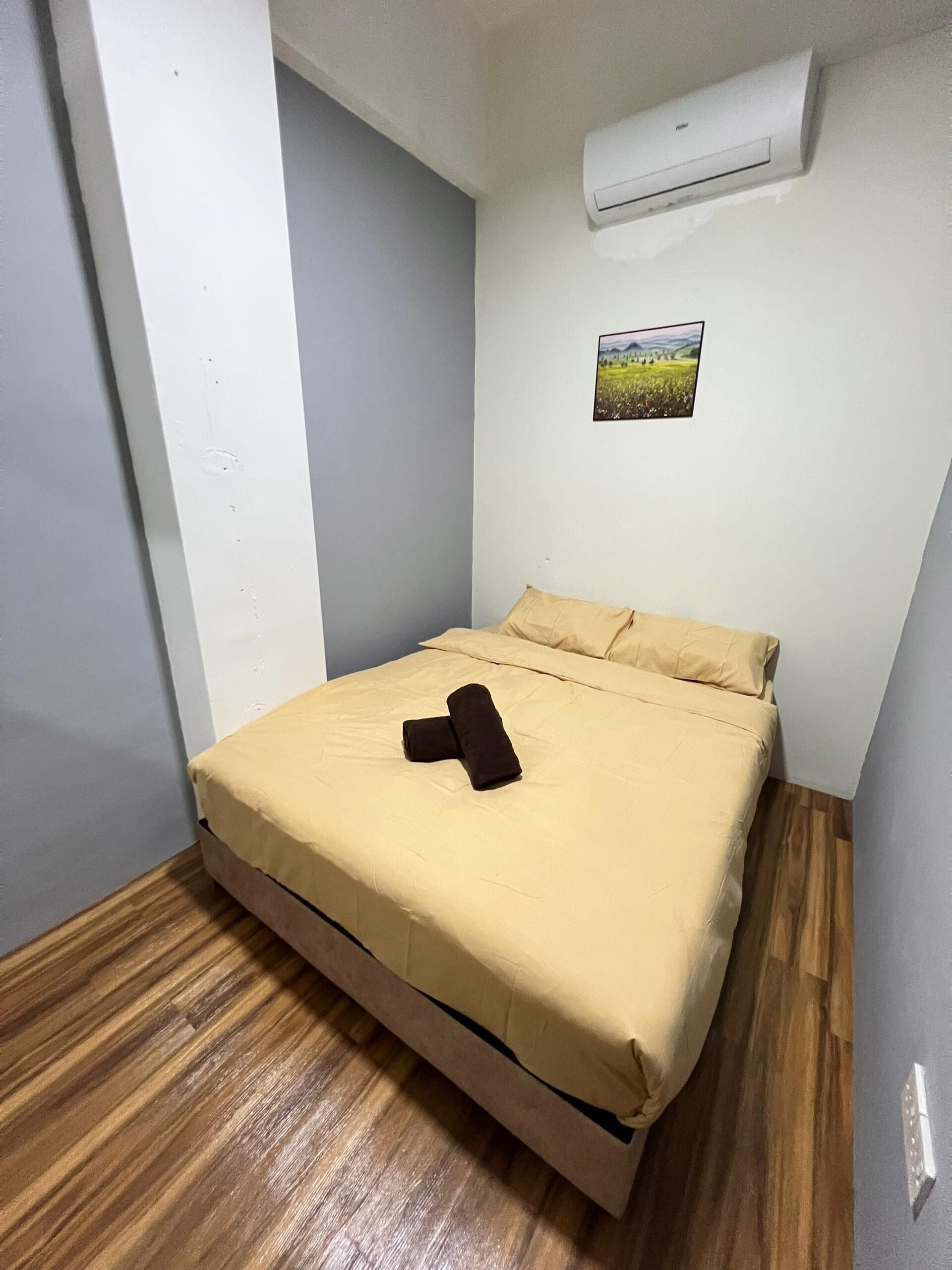 A small, single-bed room with a wooden floor, a beige bedspread, and a brown towel on the bed. There is a small picture on the wall and an air conditioning unit above the bed.