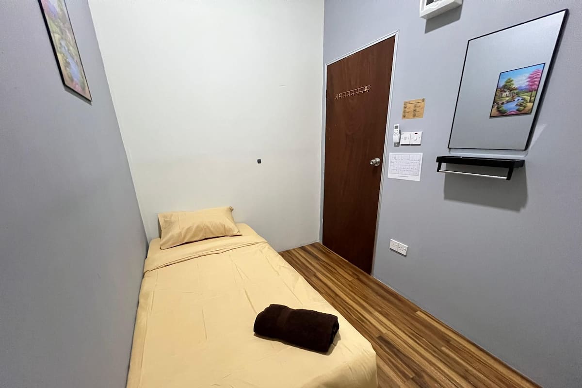Ein kleines Zimmer mit einem Einzelbett, Holzboden, einer beigen Tagesdecke und einem braunen Handtuch auf dem Bett. Eine Holztür mit Metallgriff sowie ein kleines Regal mit Bilderrahmen an der Wand.