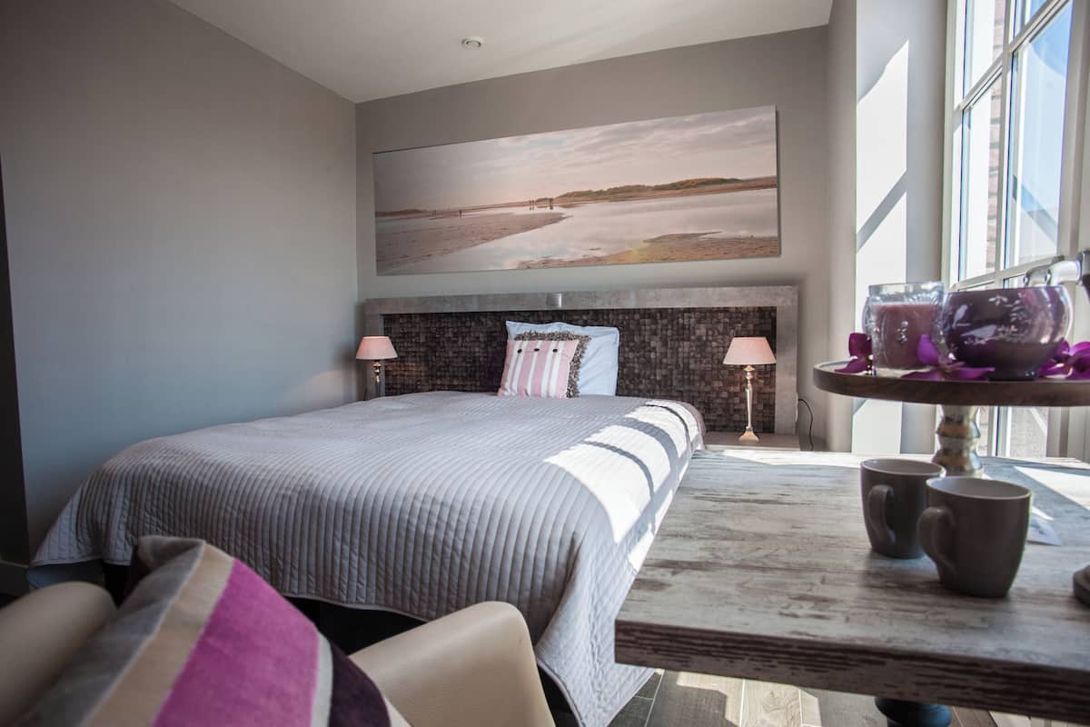 A bedroom with a large bed, a headboard with a textured design, two bedside lamps, a view of the beach through a window, and a table with two mugs.