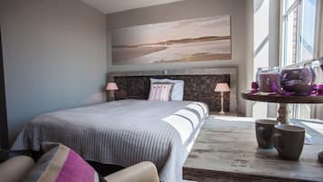 A bedroom with a large bed, a headboard with a textured design, two bedside lamps, a view of the beach through a window, and a table with two mugs.