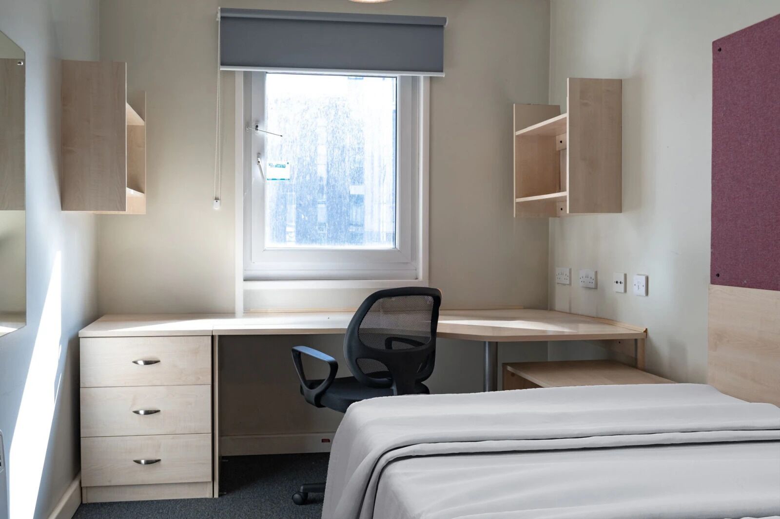 A bedroom with a single bed, a desk with a chair, a window, and built-in shelving.