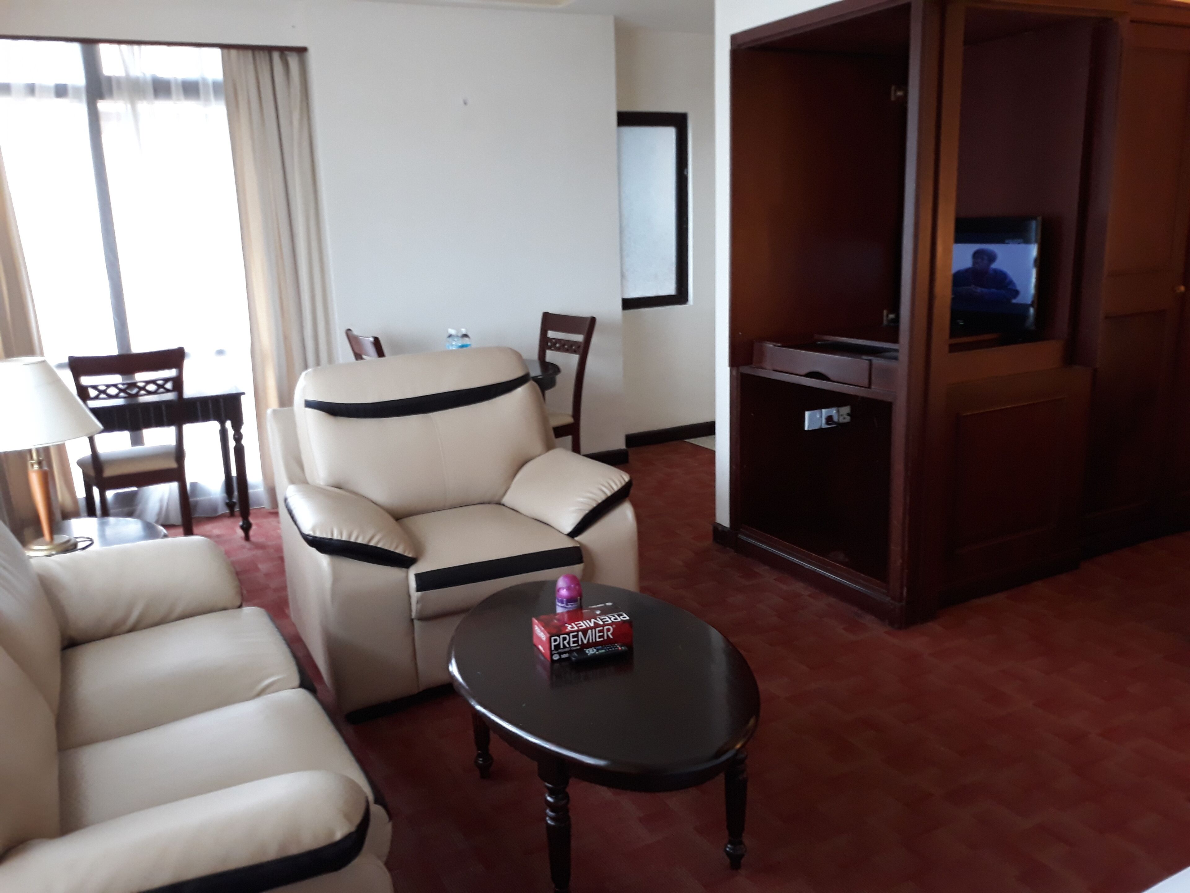 A hotel room with a beige armchair, a sofa, a small table with a box, a television, and a wooden cabinet.
