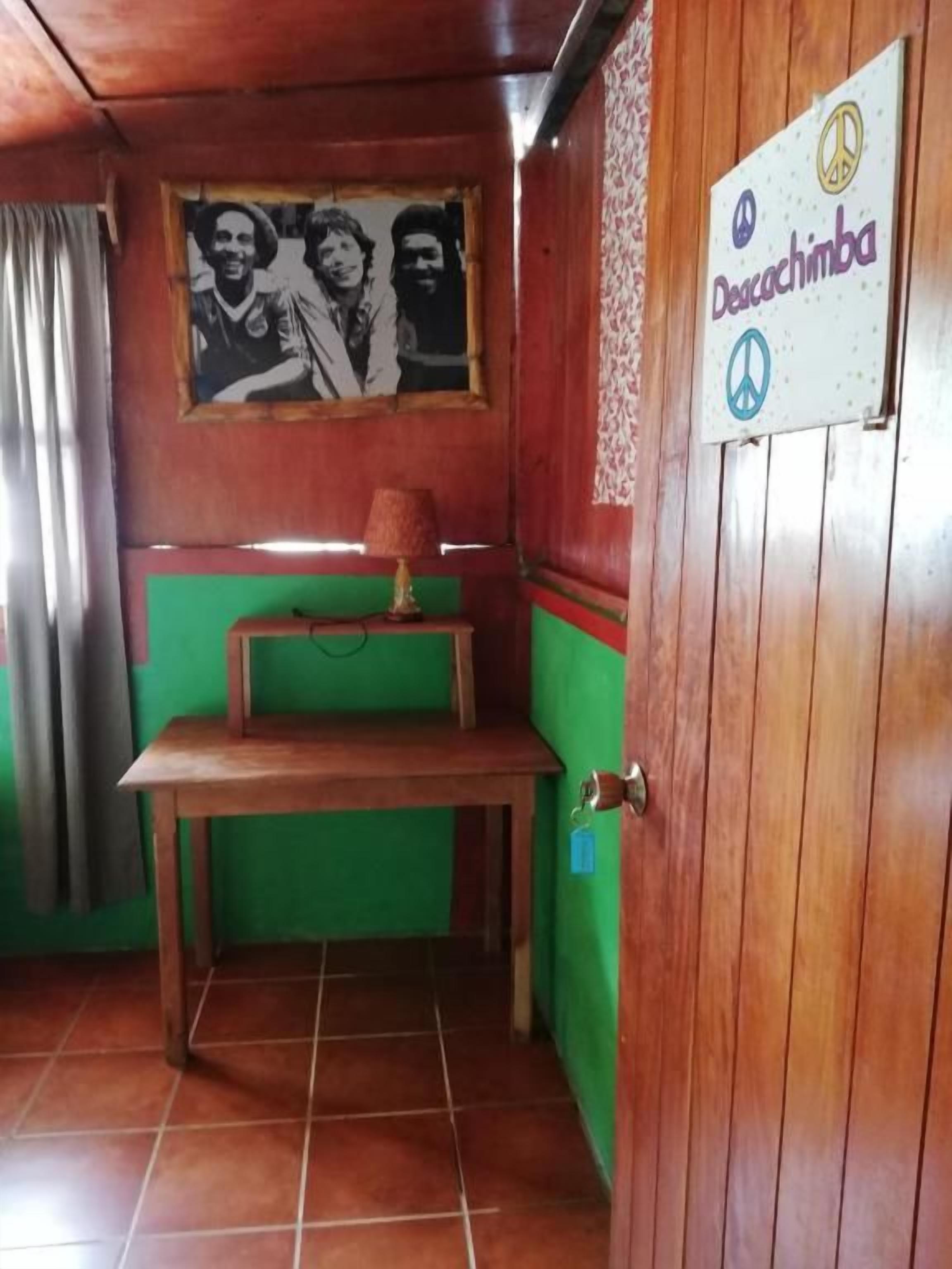A room with a wooden door, a green wall, a framed photograph, a small table with a lamp, and a sign with a peace symbol and text.