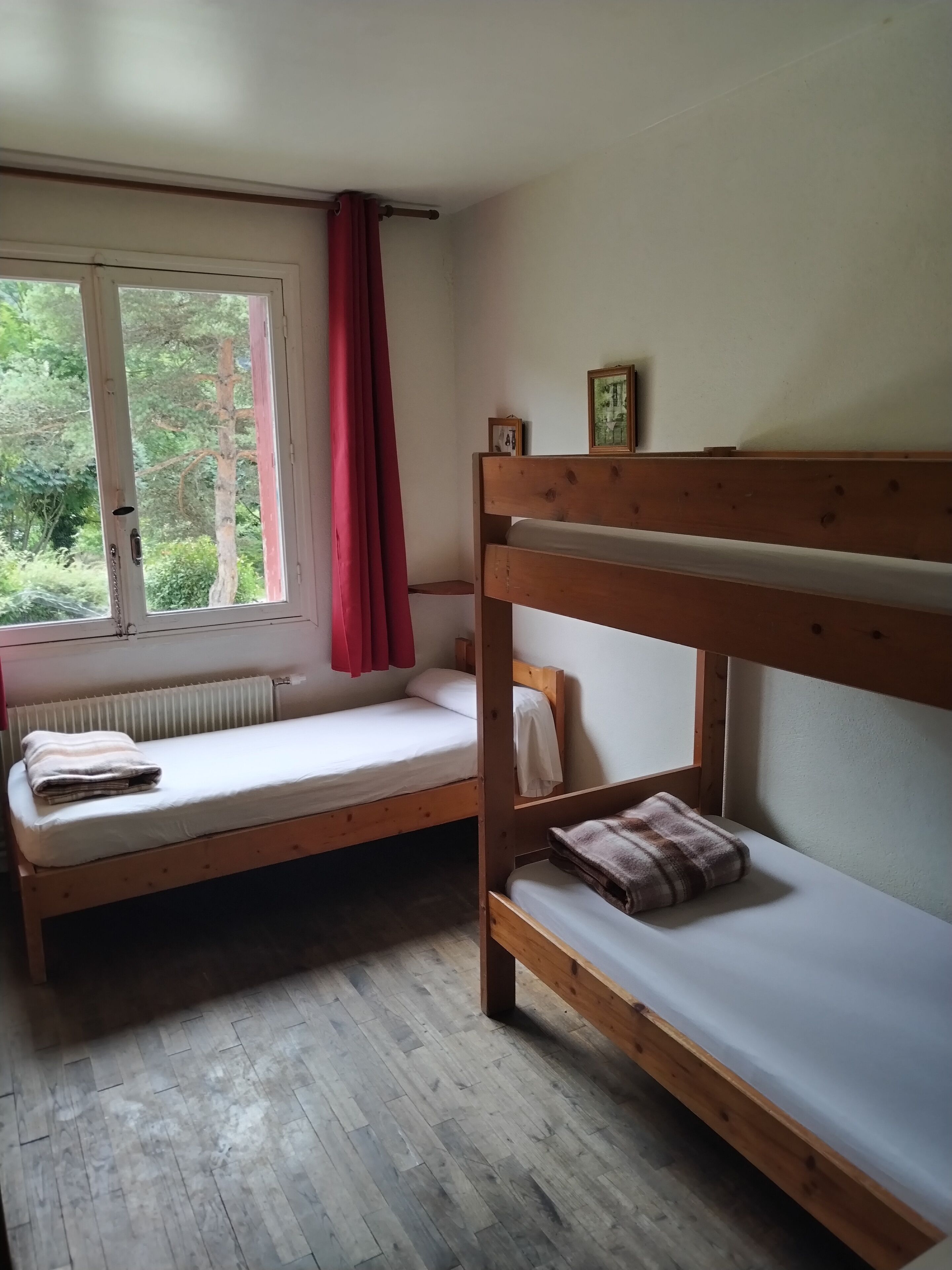 Um quarto tipo dormitório com duas beliches, uma janela com vista para árvores e um radiador sob a janela.