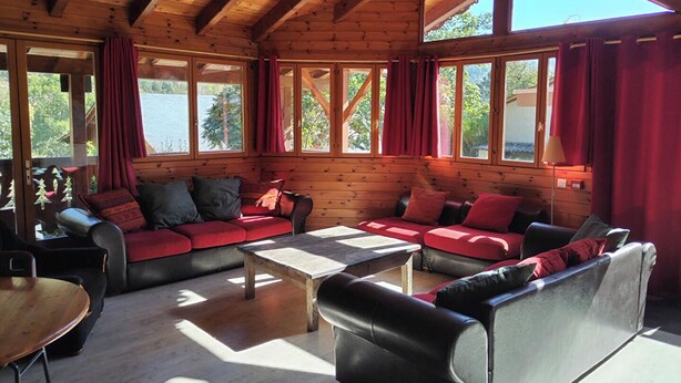Interior acogedor de una cabaña con paredes y techo de madera, un conjunto de sofás, una mesa de centro y ventanas grandes con cortinas.