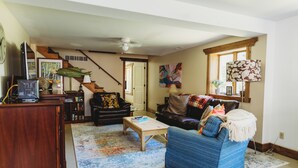 A living room with a wooden coffee table, a blue sofa, a bookshelf, and a staircase leading to another level. - Cozy Nook by Lake MI&Dunes with Private Hot Tub! (Harbert)