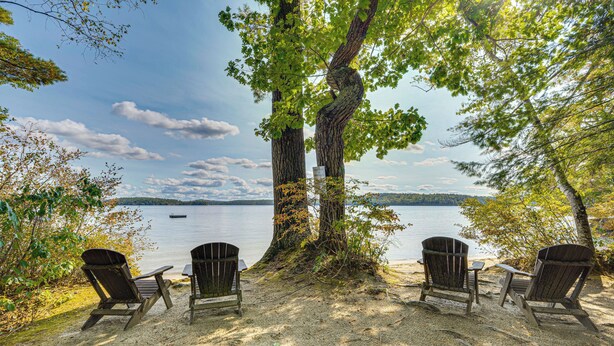 Una spiaggia con due poltrone Adirondack rivolte verso un lago, circondata da alberi.