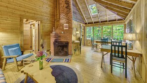 Interno accogliente di una baita rustica con camino, mobili in legno e una grande finestra vista su un paesaggio verde.