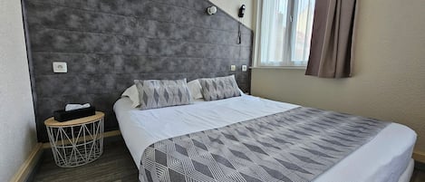 A modern hotel room with a large bed, a grey headboard, a bedside table with a tissue box, and a small trash bin.