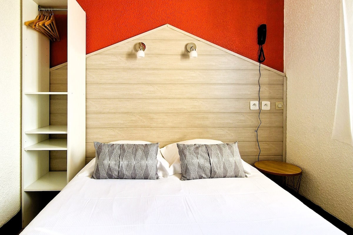 A bedroom with a bed, two patterned pillows, a wooden headboard, a white shelf unit, and a small wooden table.