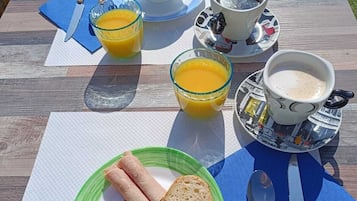 A breakfast setup on an outdoor table with a croissant, bread, and a plate of ham and cheese. There are two glasses of orange juice and a cup of coffee.