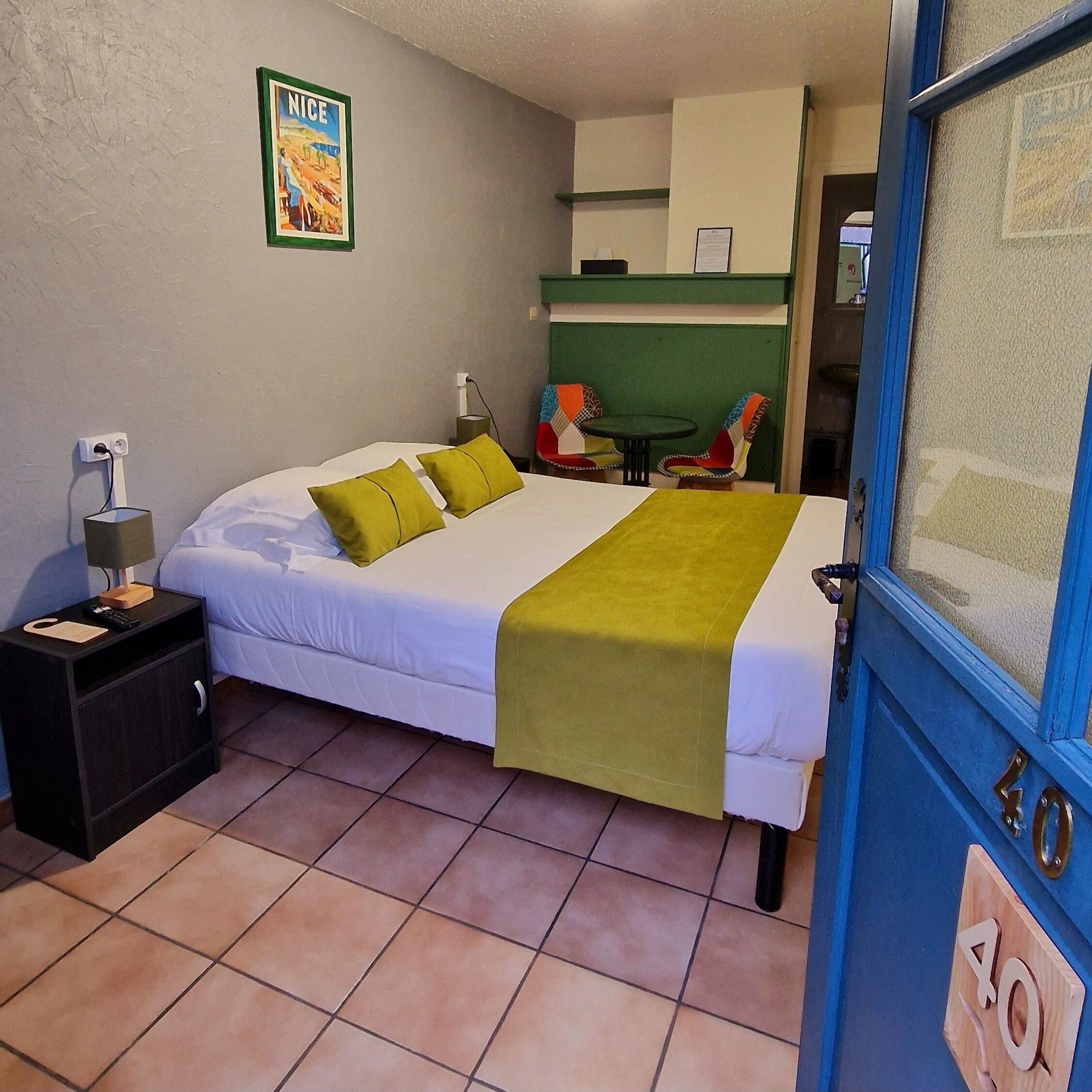 Une chambre d’hôtel avec un lit, une table de chevet, une table avec des chaises, un mur vert et une porte portant un numéro.