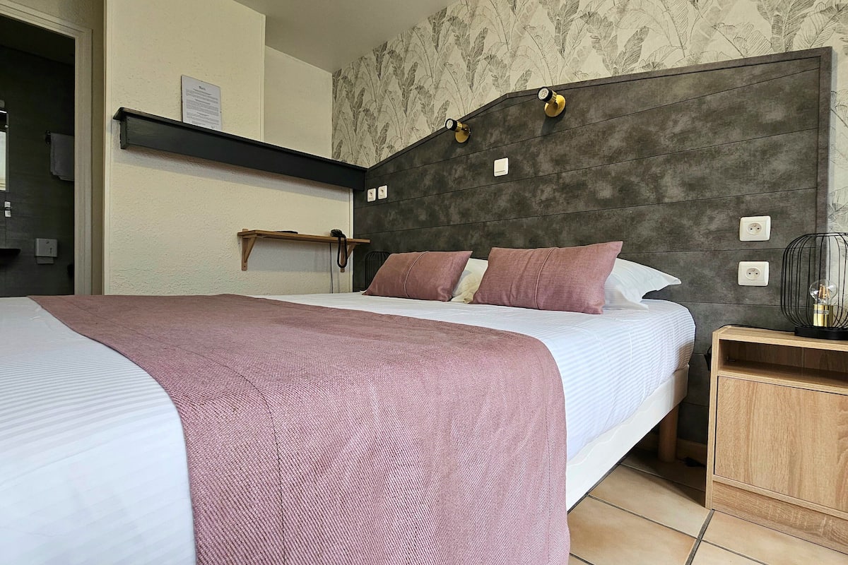 A bedroom with a bed, a bedside table, a nightstand, a wall-mounted shelf, and a wall clock.