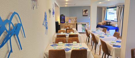 A dining area with tables set for meals, chairs, a kitchen area, and a window with curtains.