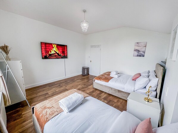 A modern hotel room with a wooden floor, a bed with white bedding, a second bed with a brown headboard, a television displaying a fireplace, a chandelier, and a small trash bin labeled "laundry". - Apartment-sleeps6-parking-internet (London)