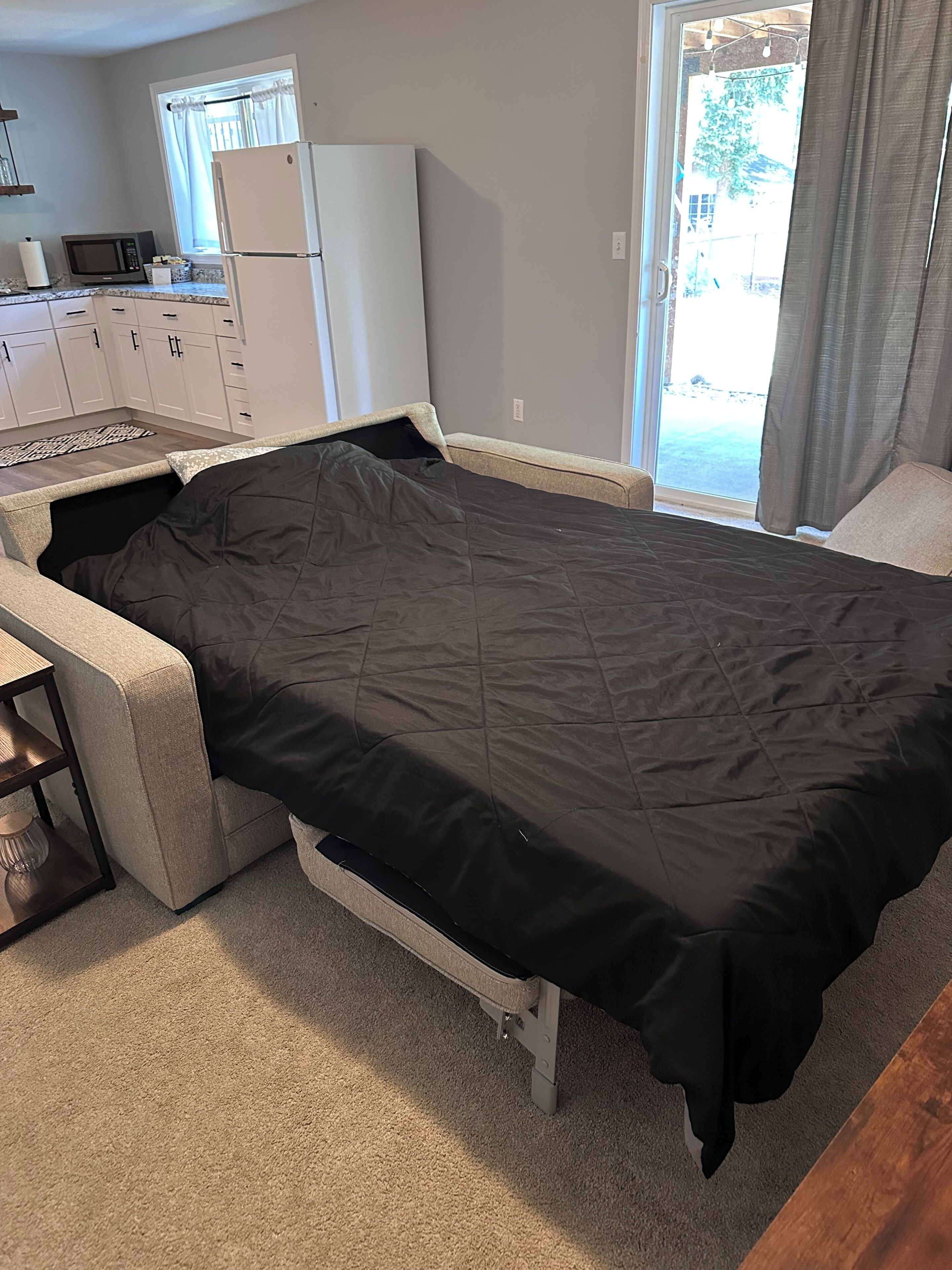 A single bed with a black mattress in a room with a kitchenette, a window with curtains, and a view of the outdoors.