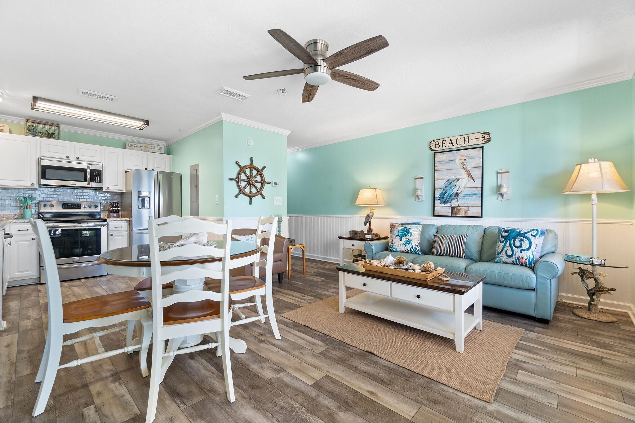 A kitchen with white cabinets, a microwave, and a refrigerator. There is a dining table with chairs and a ceiling fan. The living room has a blue couch, a coffee table, and a lamp.