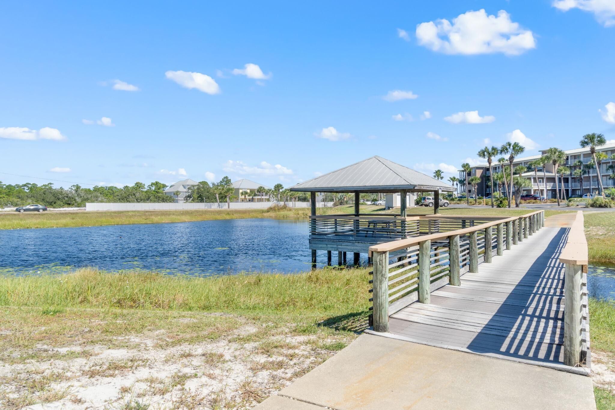 A wooden boardwalk leading to a gazebo over a body of water, with a beach and buildings in the background.