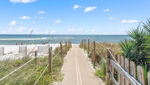 Ein hölzerner Steg führt zu einem Strand, im Hintergrund sind Menschen zu sehen.