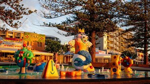 Una fontanella con diverse sculture colorate, un edificio con il cartello di un ristorante thailandese e un albero sullo sfondo.
