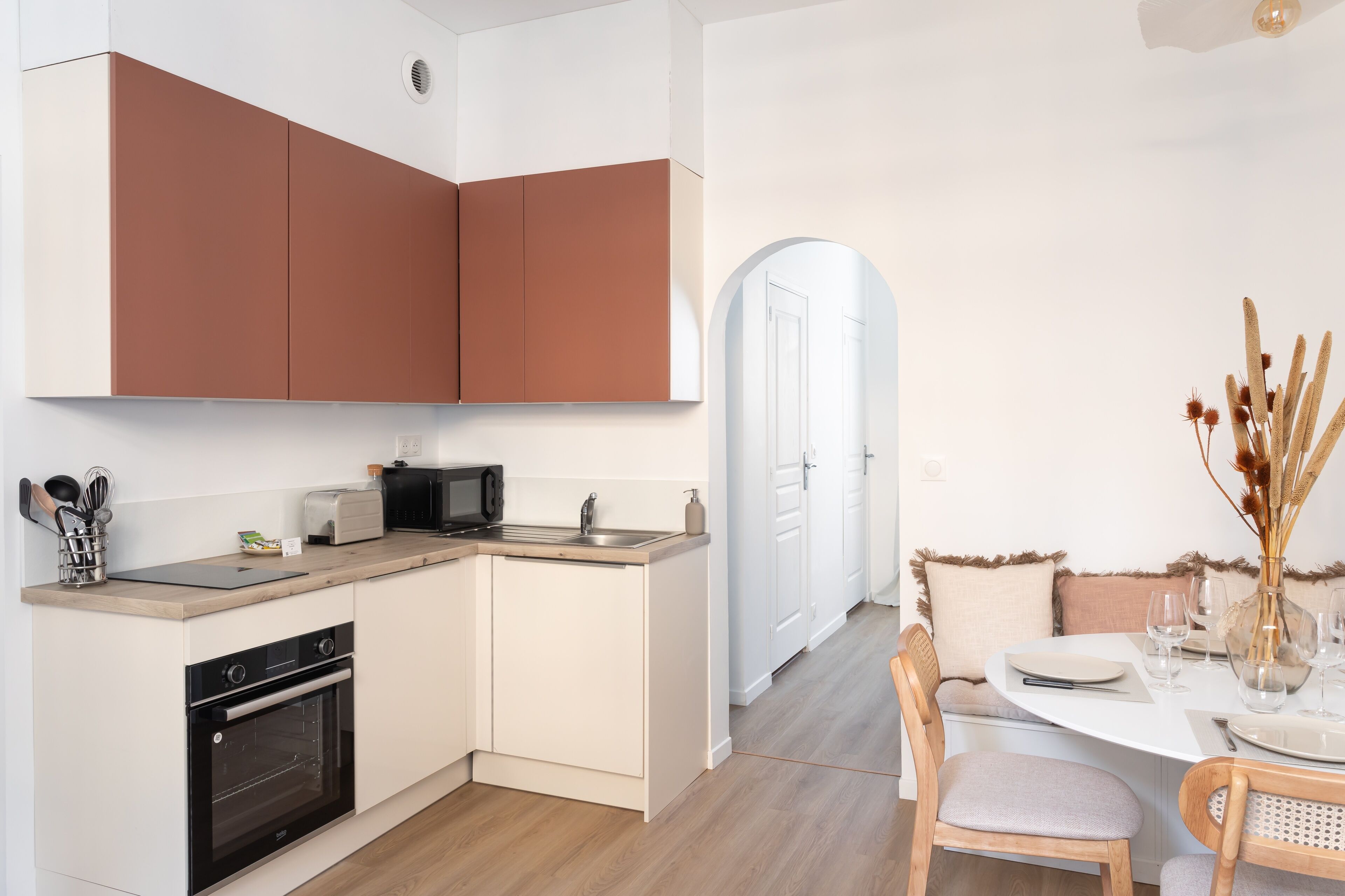 A modern kitchen with white and brown cabinets, a microwave, toaster, and a sink. There's a dining area with a table set for two, chairs, and a vase with dried flowers.