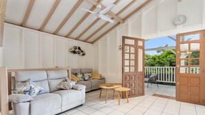 A spacious living area with a sofa, a coffee table, and a dining table. There is a wooden ceiling with exposed beams and a ceiling fan. A door leads to an outdoor patio with a chair and a table.