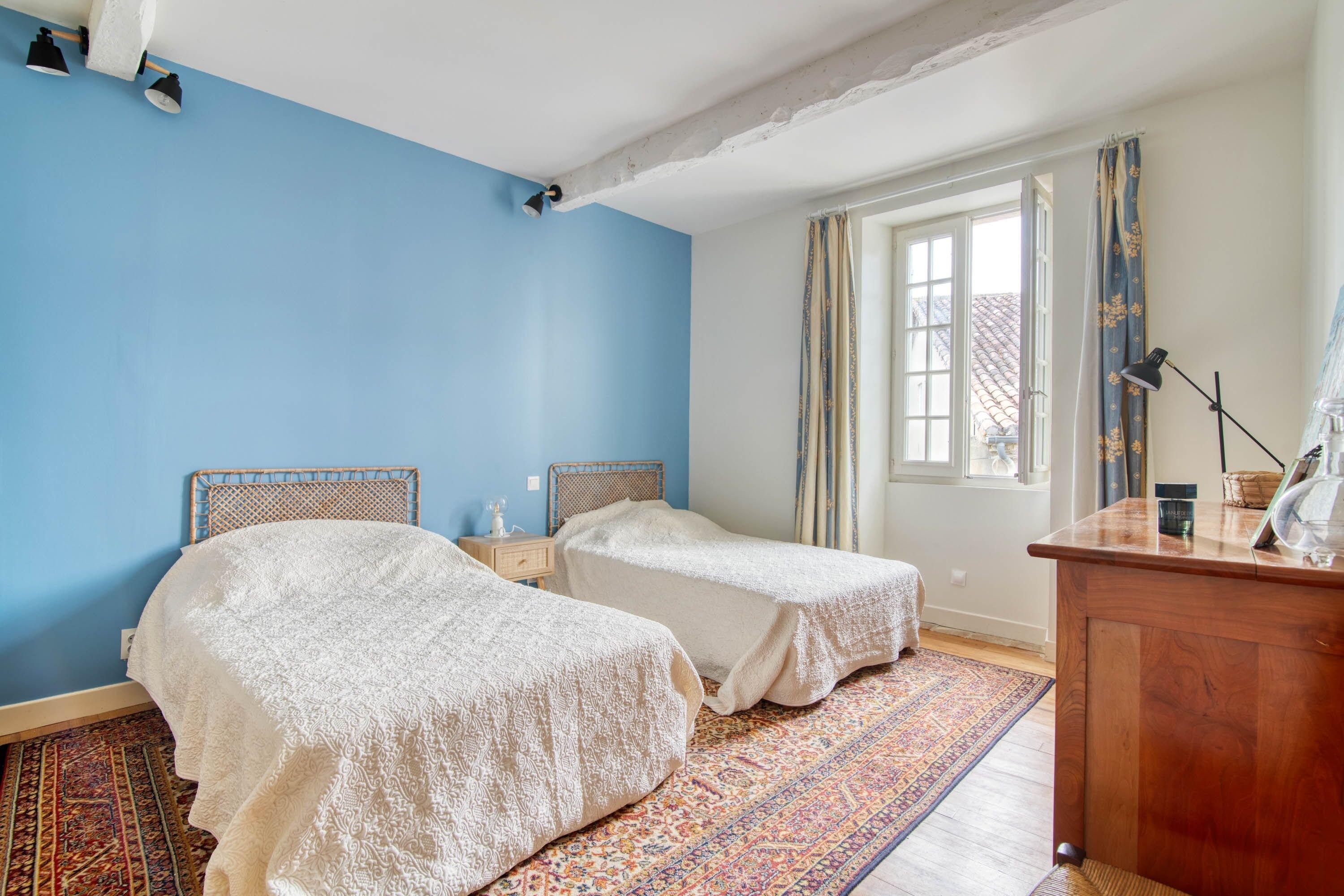 Una habitación con dos camas, un aparador de madera, una ventana con cortinas y una alfombra estampada.
