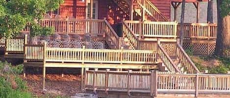 A wooden cabin with multiple decks and a small staircase, surrounded by trees and a body of water.