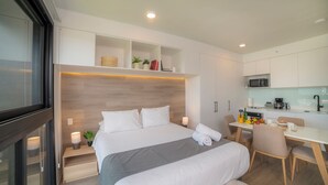 Une chambre d’hôtel moderne dotée d’un grand lit, de tables de chevet, d’un bureau avec des chaises, d’une kitchenette et d’étagères intégrées.