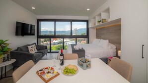 Une chambre d’hôtel moderne dotée d’une grande fenêtre, d’un lit, d’un canapé, d’une table à manger avec de la nourriture et des boissons, et d’un téléviseur.