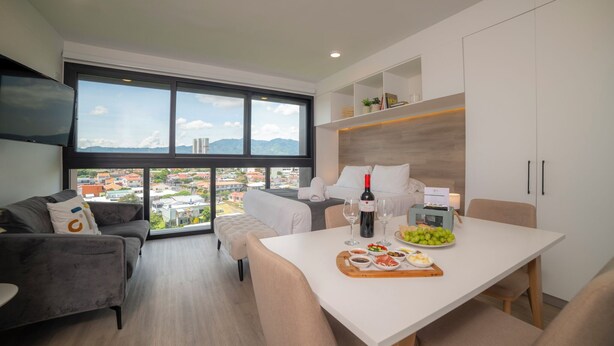 Une chambre d’hôtel moderne dotée d’une grande fenêtre donnant sur la ville, d’un coin repas avec une table dressée pour deux personnes, et d’un espace salon confortable avec un canapé et un pouf.