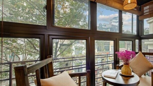 A room with large windows, a wooden chair with cushions, a small round table with a book and a vase of flowers.