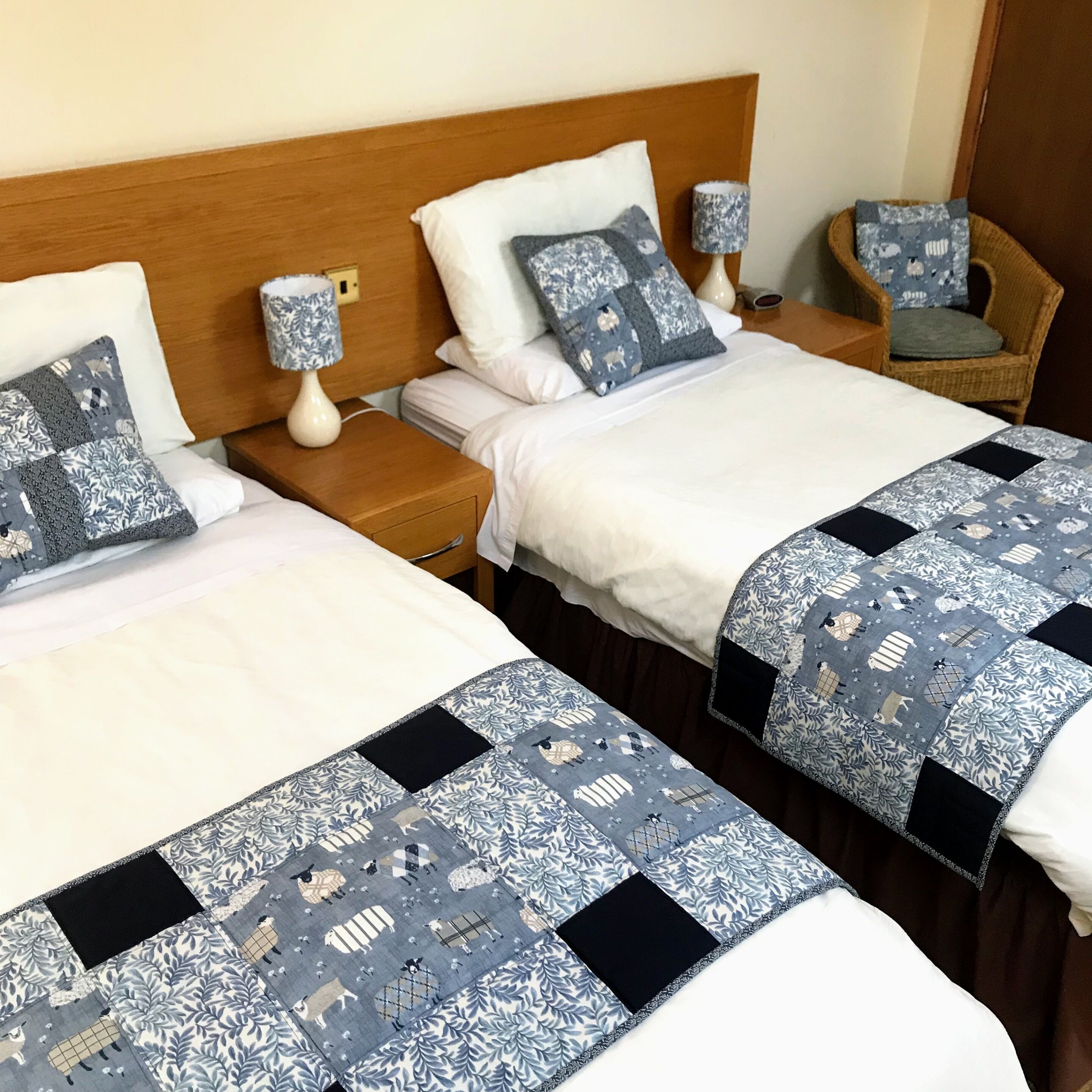 A hotel room with two beds, each with a blue and white patterned bedspread and matching pillows. There are two bedside tables with lamps and a wicker chair.
