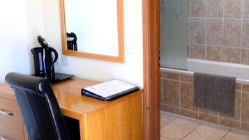 A room with a wooden desk, a black chair, a mirror, a coffee maker, and a tiled bathroom with a towel hanging on the wall.