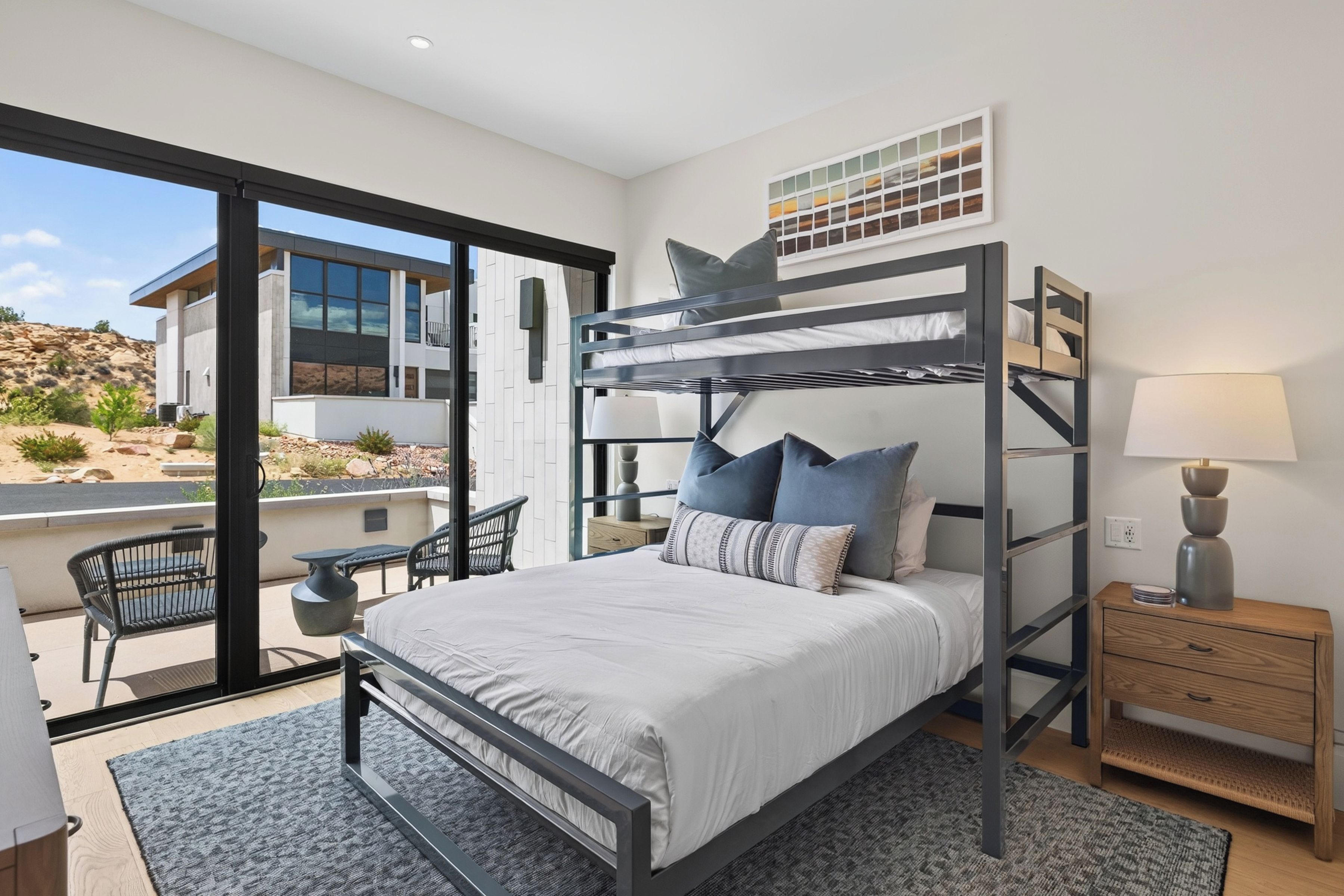 A modern bedroom with a bunk bed, a desk, and a chair, featuring a large window and a view of a building.