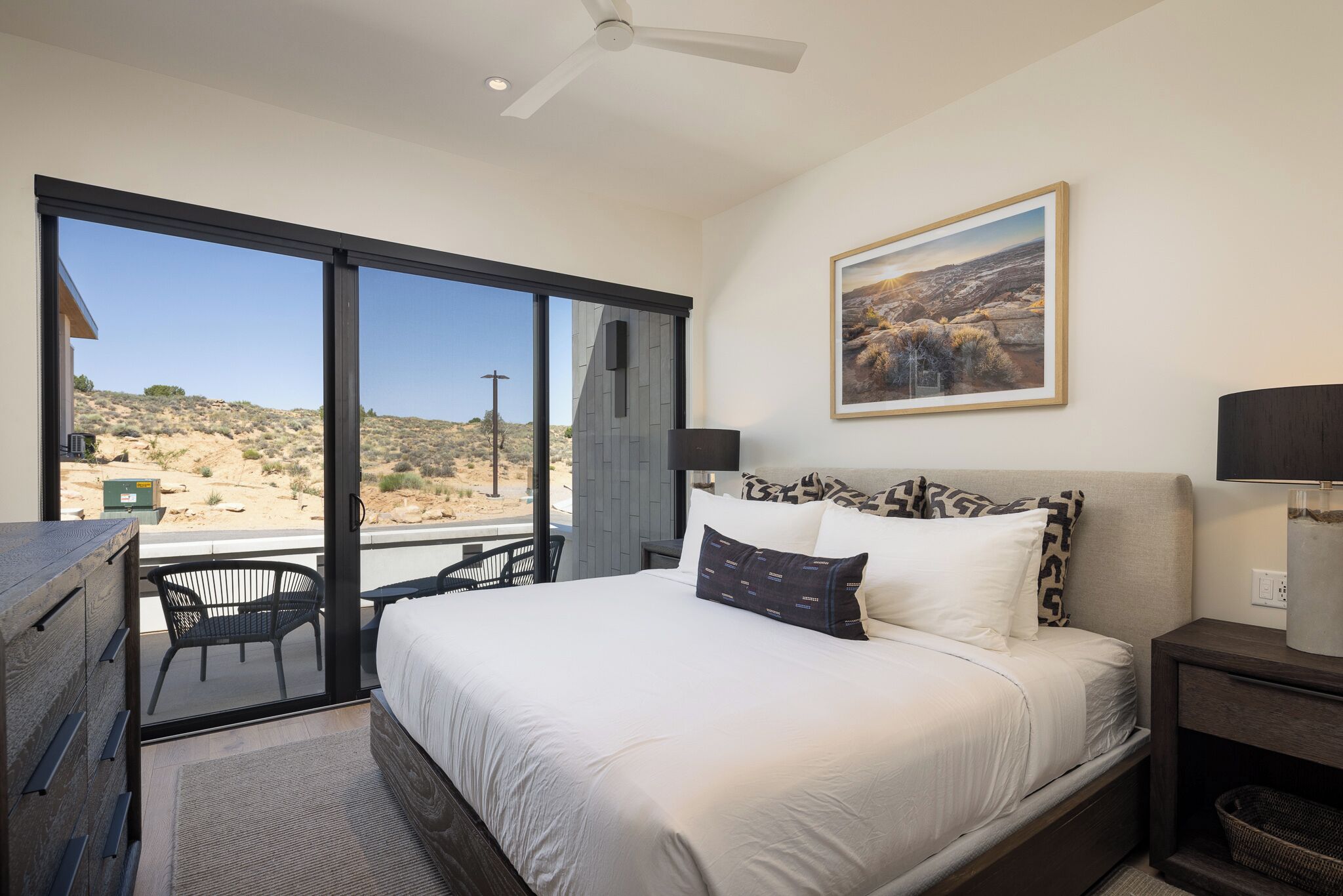 A bedroom with a bed, a nightstand, a lamp, a painting, and a view of the desert through large windows.