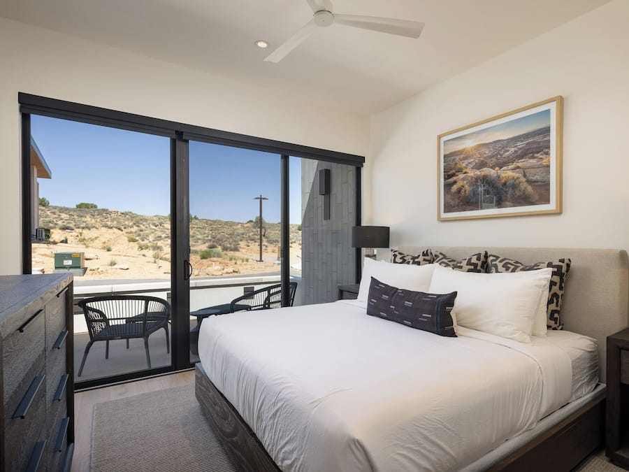 A bedroom with a bed, a nightstand, a lamp, a painting, and a view of the desert through large windows.
