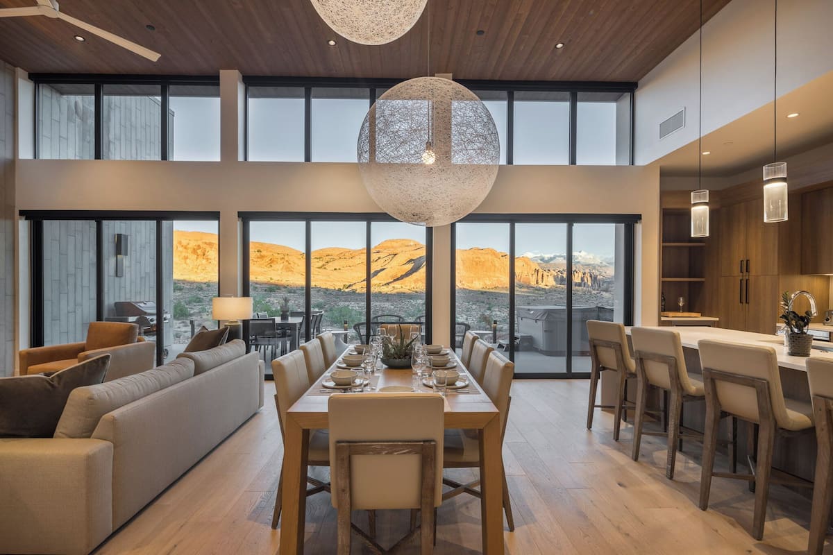Uma sala de jantar espaçosa com uma grande mesa posta para seis pessoas, um sofá e uma área de cozinha com armários de madeira e iluminação pendente.