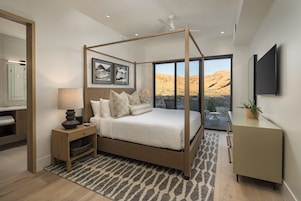 Un dormitorio con cama con dosel, una mesita de noche con lámpara, un ventanal con vistas al desierto y un televisor de pantalla plana montado en la pared.