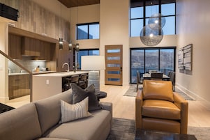 A spacious living area with a sofa, a leather chair, and a dining table. There is a kitchen with wooden cabinets and a large window overlooking a mountainous landscape.