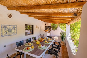A dining area with a wooden table set for breakfast, surrounded by chairs, a hanging light fixture, and a view of greenery outside. - Villa Alice (Albufeira)