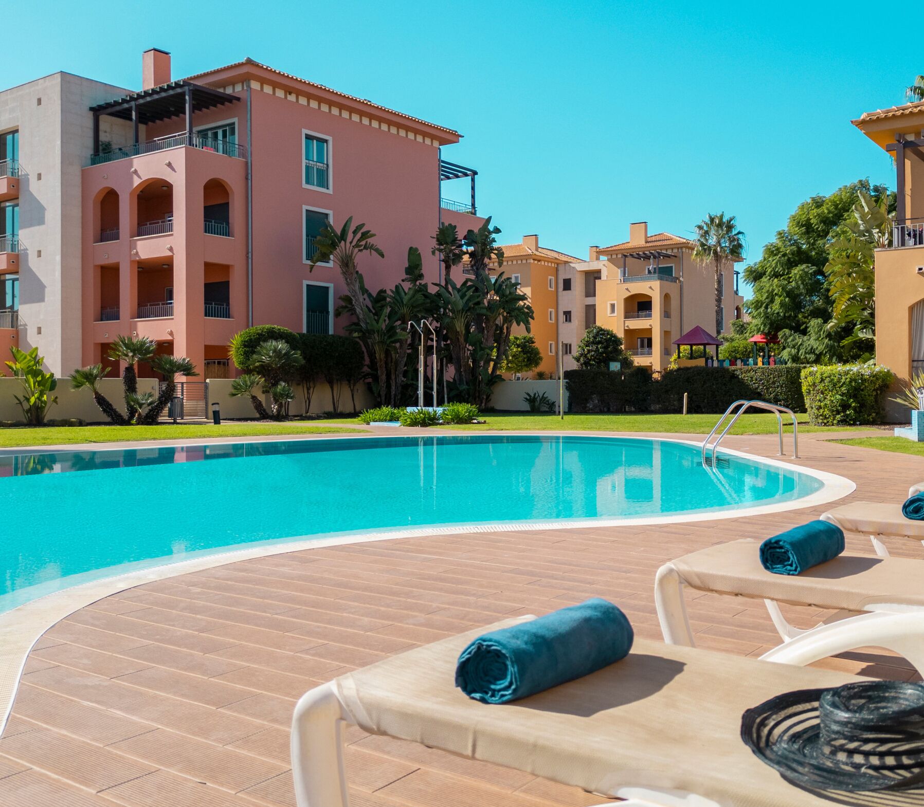 A swimming pool with lounge chairs, a clear blue sky, and multi-story buildings with balconies.