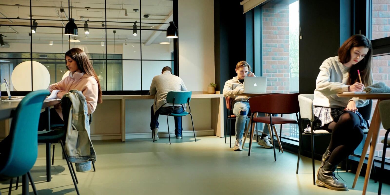 A modern office space with multiple workstations, a large window with a brick wall, and hanging pendant lights.