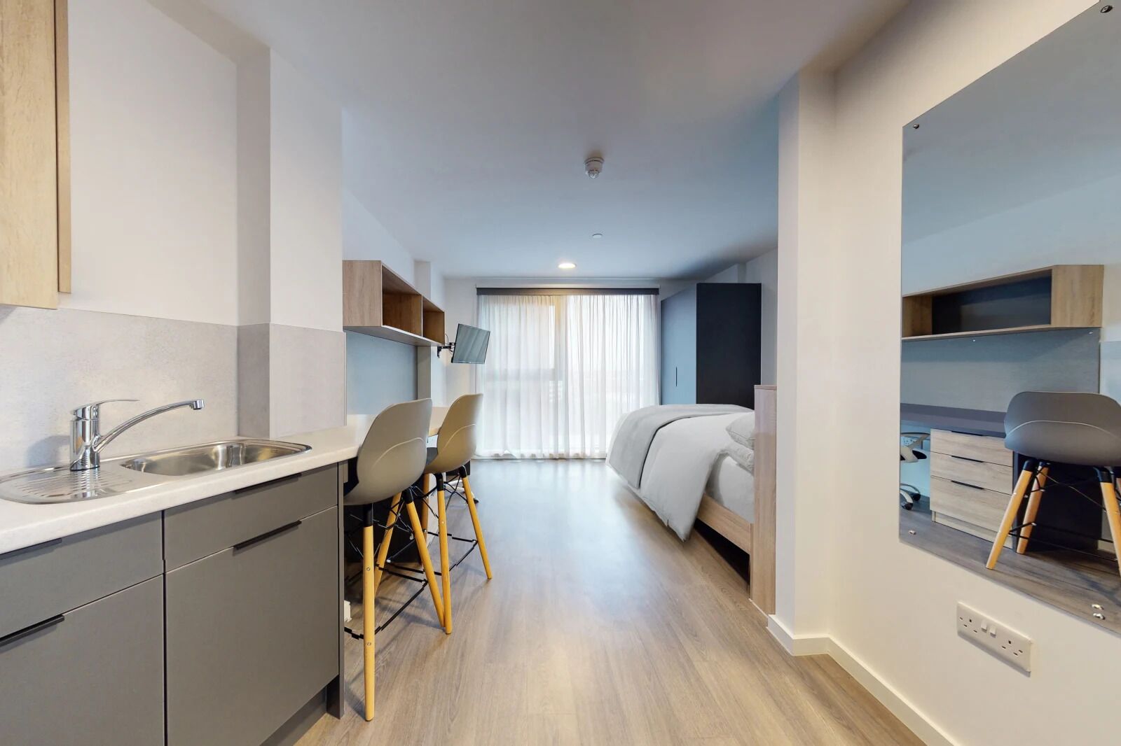 A modern, compact hotel room with a kitchenette, a bed, a desk, and a chair.