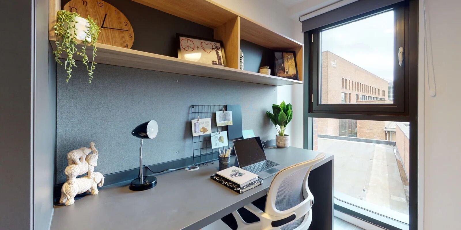 A modern office desk with a laptop, a desk lamp, and a small statue. There are shelves above the desk holding decorative items and a plant.