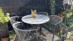 Une terrasse avec une table ronde en métal et quatre chaises en osier, entourée de plantes en pot et d’une clôture noire.
