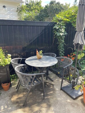A patio with a round metal table and four wicker chairs, surrounded by potted plants and a black fence.