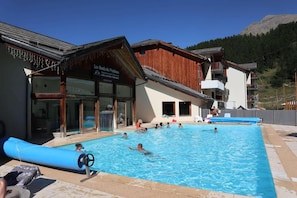 A swimming pool area with people swimming and lounging, a wooden building with a sign, and a mountain in the background.