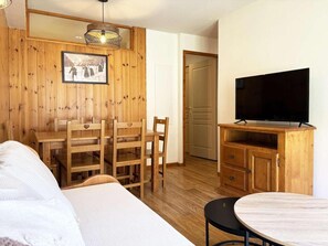 A cozy room with a wooden wall, a dining table with chairs, a TV on a wooden cabinet, and a bed.